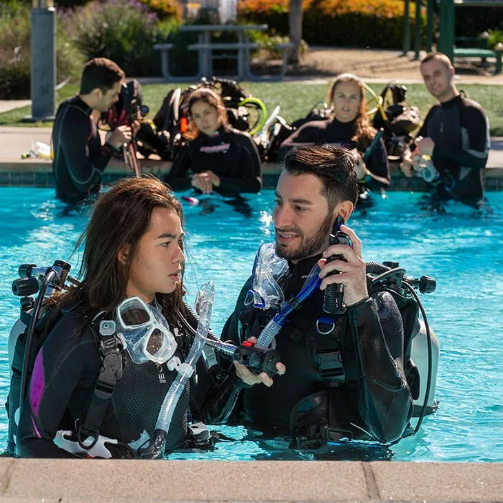 Instructora y alumno practican en la piscina con equipo completo mientras el grupo observa