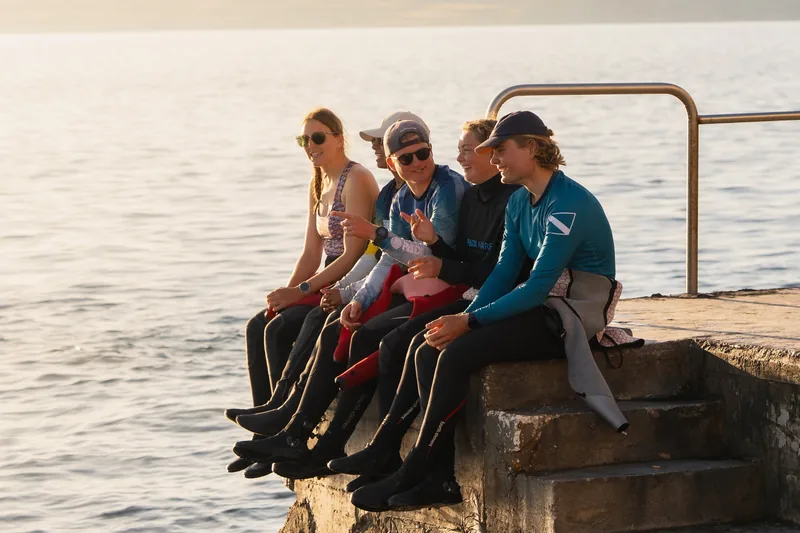 Cuatro buceadores con neopreno sentados en un escalón del muelle al atardecer