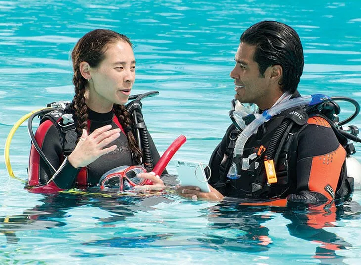 Instructora explica con gestos a un alumno dentro de una piscina turquesa con regulador