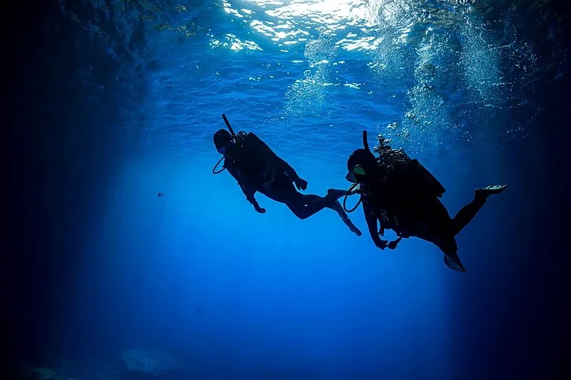 Dos buceadores a contraluz sobre el azul con burbujas subiendo hacia la luz