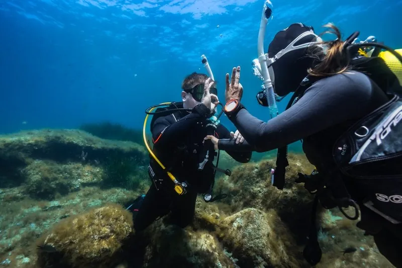 Instructor y alumno se saludan con la mano bajo el agua sobre fondo rocoso
