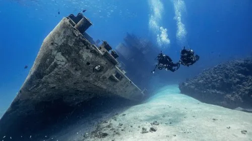 Casco de un barco hundido sobre arena clara con dos buceadores a su lado