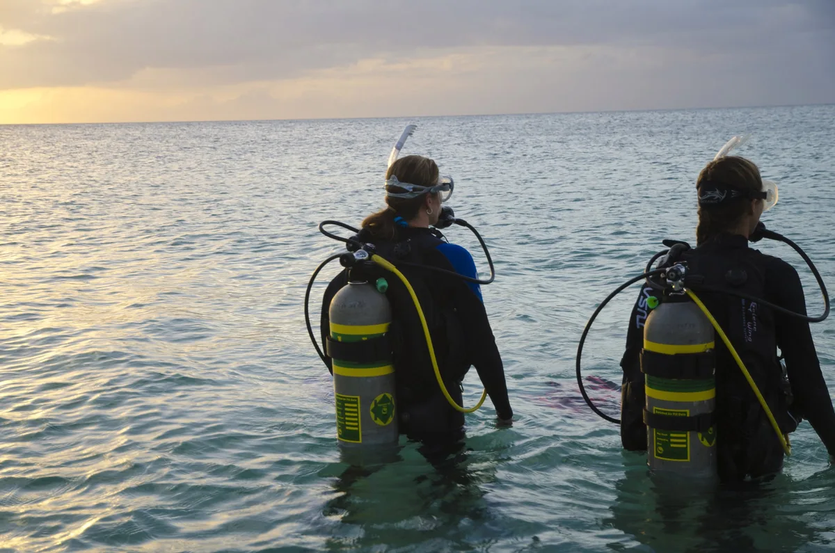 Dos buceadores con botellas amarillas entran al mar de espaldas al atardecer