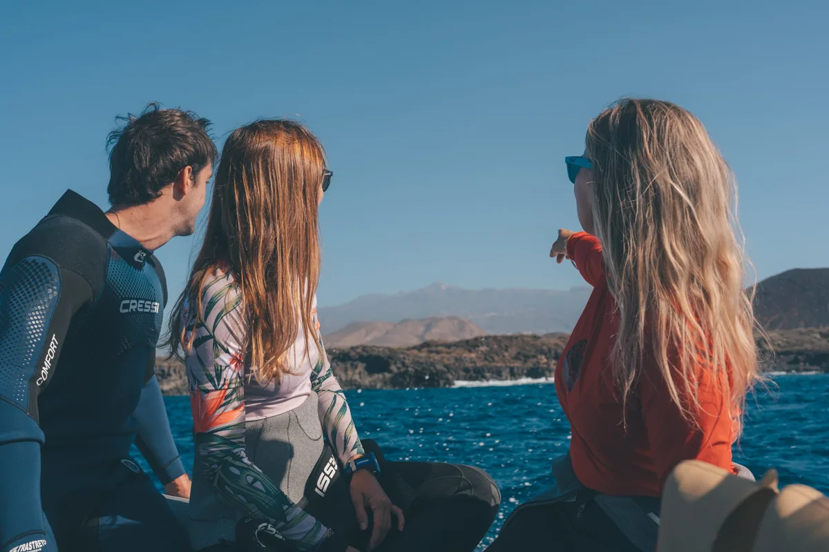 Tres personas con neopreno miran el horizonte desde un barco antes de bucear