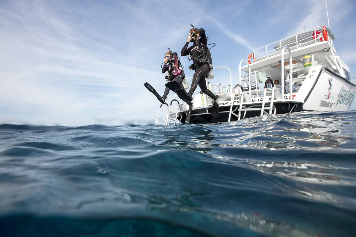 Dos buceadores esperan en la plataforma de popa de un barco blanco