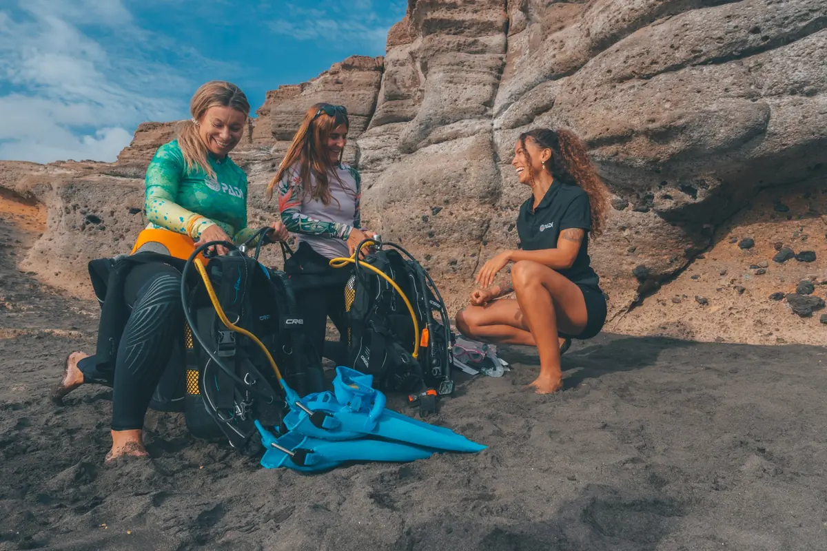 Tres buceadoras preparan sus equipos sobre rocas oscuras junto al mar