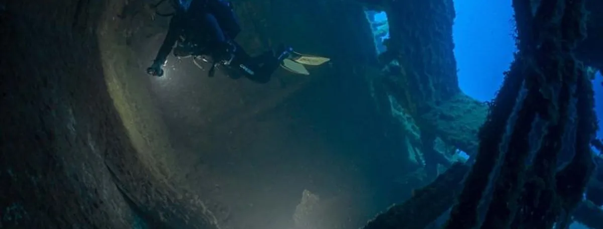 Interior oscuro de un pecio hundido con luz azul tenue y un buceador lejano