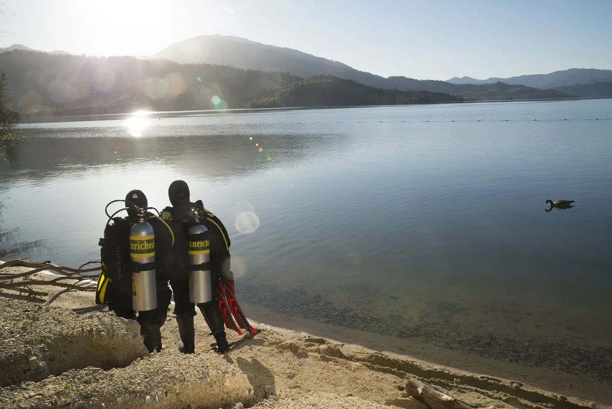 Dos buceadores de espaldas con botellas amarillas ante un lago al amanecer