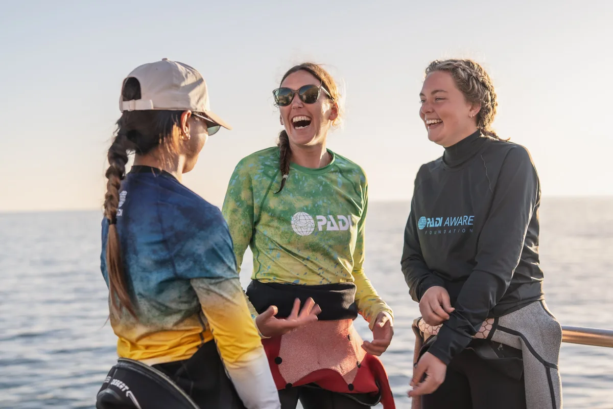 Tres instructoras jóvenes ríen en la cubierta de un barco al atardecer